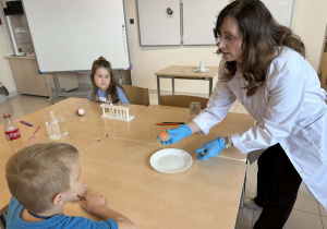 od chemii trzyma jajko bez skorupki pokazując je dzieciom siedzącym przy stoliku