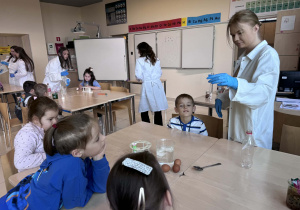 uczenica trzymając menzurkę pokazuję dzieciom rakcję zachodzącą w połaczeniu skorupki jajka i octu