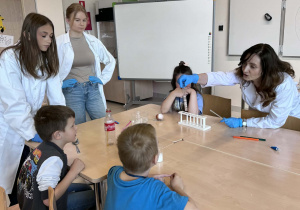 pani od chemii trzymając menzurkę z skorupkami jajek pokazuję dzieciom i uczenicom jaka reakcja zachodzi w połączeniu z octem