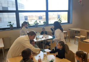 dzieci siedzą przy stolikach i wykonują doświadczenia z jajkiem przy pomocy uczenic liceum i nauczycielki chemii