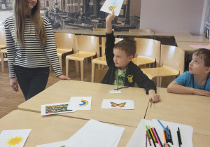 na stoliku leża porozrzucane obrazki, jedenk chłopiec trzyma obrazke w górze, obok niego stoi uczenica liceum