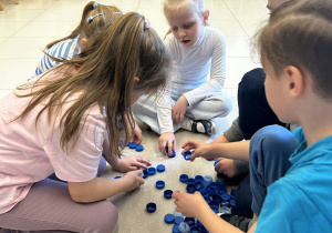 Dzieci pracują w grupie tworząc figury geometryczne z niebieskich zakrętek