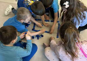 Dzieci pracują w grupie tworząc figury geometryczne z niebieskich zakrętek