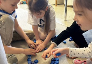 Dzieci pracują w grupie tworząc figury geometryczne z niebieskich zakrętek