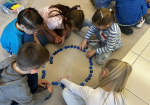 Dzieci pracują w grupie tworząc figury geometryczne z niebieskich zakrętek