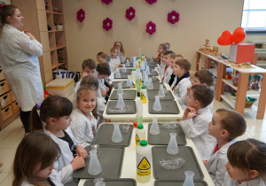 dzieci siedzą przy stolikach i słuchają poleceń pani prowadzącej warsztaty