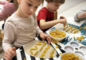Dzieci wyklejają papierowe pisanki różnymi rodzajami makaronu.