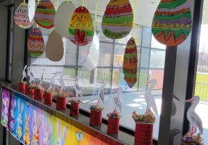 Gotowe pisanki wiszą w oknie nad papierowymi bocianami w gniazdach.