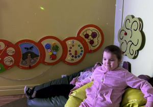 Dziewczynka odpoczywa na wygodnej pufie w sali z sensorycznymi panelami ściennymi i świetlnymi efektami na ścianie.