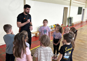 Nauczyciel rozmawia z grupą dzieci na sali gimnastycznej, przygotowując je do kolejnego ćwiczenia.