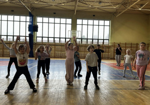 Dzieci wykonują pajacyki na środku dużej hali sportowej.