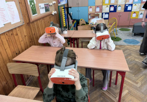 Troje dzieci siedzących w ławkach korzysta z gogli do rzeczywistości wirtualnej