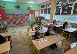 Grupa dzieci w kolorowej sali lekcyjnej bierze udział w zabawie dydaktycznej, podnosząc do góry kartoniki z rysunkami owoców.