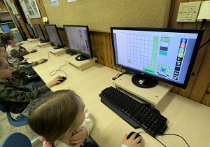 Sześciolatki w pracowni komputerowej rozwiązują logiczne zadania i układanki na monitorach stacjonarnych.