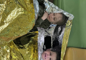 Dwóch chłopców leży na podłodze przykrytych złotą folią życia (termiczną) podczas praktycznych ćwiczeń z pierwszej pomocy.