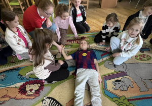 Grupa dzieci wraz z panią otacza kolegę leżącego na dywanie, ucząc się sprawdzania oddechu i tętna u poszkodowanego.