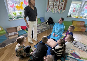 Nauczycielka prowadzi instruktaż pierwszej pomocy dla grupy dzieci siedzących na dywanie wokół fantoma medycznego.