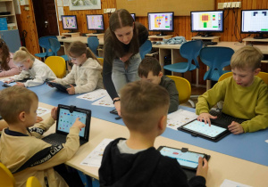 Grupa dzieci w pracowni komputerowej korzysta z tabletów, wykonując zadania edukacyjne pod nadzorem nauczycielki.