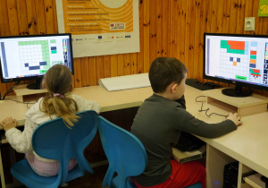 Dwoje dzieci siedzi przy stanowiskach komputerowych, rozwiązując na monitorach kolorowe zagadki logiczne oparte na siatce pikseli.