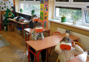 Troje dzieci siedzi w ławkach szkolnych, mając na oczach gogle VR.