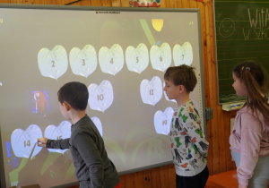 Chłopiec używa specjalnego pióra na tablicy interaktywnej, rozwiązując zadanie matematyczne polegające na łączeniu ponumerowanych elementów w kształcie liści.