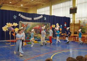 Grupa dzieci przebranych za Smerfy występuje na scenie w hali sportowej podczas uroczystego przedstawienia.