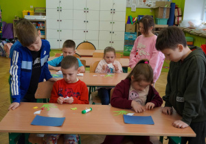 Grupa dzieci skupiona przy stolikach wykonuje prace plastyczne z kolorowego papieru i kleju.