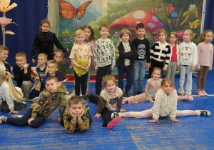 Pamiątkowe zdjęcie grupy dzieci na tle bajkowej dekoracji z motylami i muchomorem.