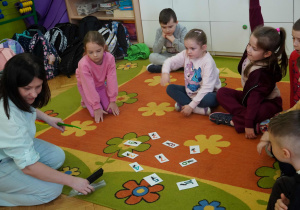 Dzieci siedzą w kręgu na kolorowym dywanie i biorą udział w edukacyjnej zabawie z wykorzystaniem kartoników z liczbami.