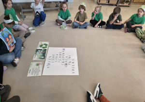 Grupa dzieci siedzi w kręgu wokół ułożonej krzyżówki i tablic informacyjnych o recyklingu.