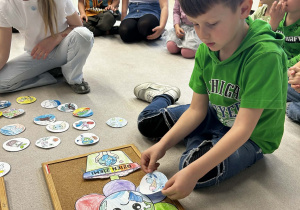 Chłopiec w zielonej bluzie dekoruje papierowy kwiatek z motywem kuli ziemskiej kolorowymi obrazkami.