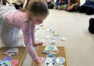 Dziewczynka w różowej bluzce przykleja małe ilustracje do dużej pracy plastycznej przedstawiającej uśmiechniętą Ziemię.