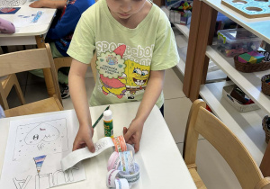 Chłopiec przy stoliku skleja papierowy łańcuch z kolorowymi rysunkami i napisami.